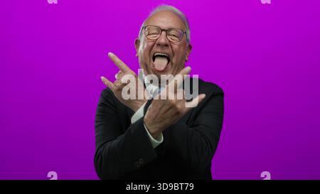 Uomo anziano in una tuta da lavoro che fa gesti di roccia con un'espressione giocosa su una parete rosa brillante sullo sfondo. Foto Stock