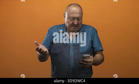 Uomo ispanico anziano che tiene lo smartphone con la mano destra e mostra un gesto del palmo aperto verso lo schermo sullo sfondo arancione dello studio; frustrazione digitale. Foto Stock
