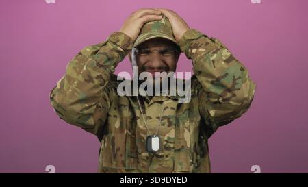 Il giovane soldato ispanico in uniforme mimetica tiene in mano le targhette per cani d'argento e preme mano sulla fronte in studio rosa; stress. Foto Stock