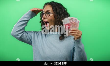 Una donna di mezza età che tiene in mano banconote in corona islandese guarda in lontananza contro una vivace parete verde, trasmettendo curiosità ed eccitazione. Foto Stock