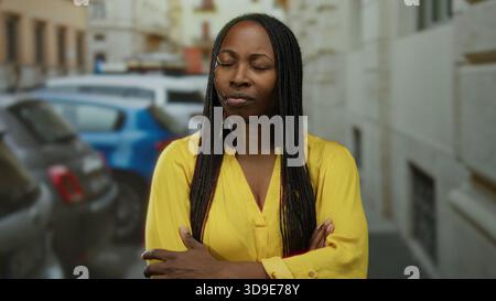 Donna in camicia gialla con capelli intrecciati si stende su strada urbana, esprimendo lo stress con gli occhi chiusi e le braccia incrociate, circondata da auto parcheggiate in un ci Foto Stock
