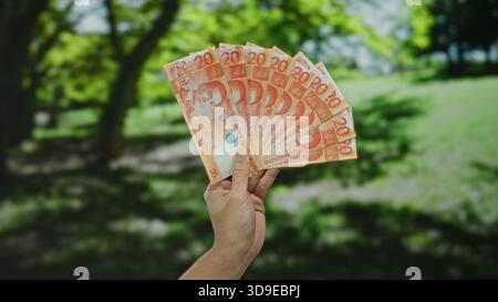 Uomo caucasico che tiene all'aperto diverse banconote in peso filippino in un parco, mettendo in evidenza un concetto di ricchezza o finanza in un ambiente naturale Foto Stock