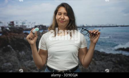 La giovane donna latina tiene gli occhiali e la custodia per lenti a contatto in mano sulla costa rocciosa per strada; scelta indecisa. Foto Stock