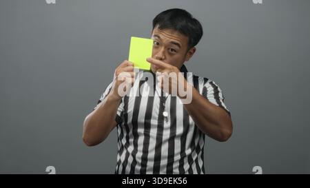 Il giovane arbitro cinese tiene il cartellino giallo e punta il dito mentre indossa una camicia a righe nello studio grigio; autorità. Foto Stock