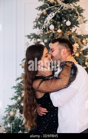 Una coppia amorevole condivide un momento tenero con un albero di Natale festivo. Sono vestiti elegantemente, irradiano felicità mentre si godono il calore dell'ho Foto Stock