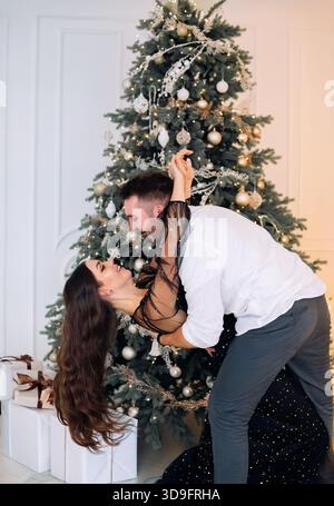 Una donna con un elegante abito nero e la sua compagna si divertono a ballare vicino a un albero di Natale splendidamente decorato con luci e ornamenti, festeggiando Foto Stock