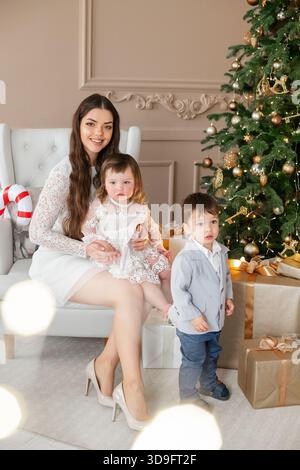 Una donna siede comodamente su una sedia bianca con due bambini piccoli accanto a lei in un soggiorno caldo. Foto Stock