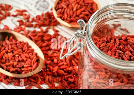 Goji secco aka. Semi di mirtillo all'interno di una bottiglia di vetro, alcuni più sfocati in ciotole di legno e versato sulla scrivania delle tavole bianche Foto Stock