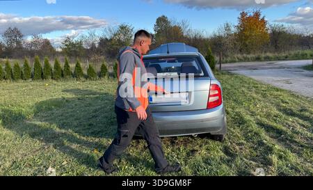 Un meccanico caucasico maschile con una giacca grigia e arancione che apre il bagagliaio di un'auto d'argento parcheggiata su una strada erbosa sotto un cielo azzurro. Foto Stock