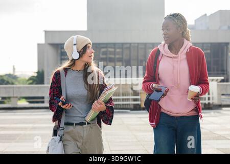 Diverse amiche che camminano attraverso il campus plaza chiacchierando con telefoni cuffie appunti bustine di caffè Foto Stock