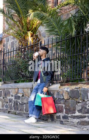 Uomo in piedi sul marciapiede della città che tiene le borse della spesa e solleva la tazza di caffè con una borsa a tracolla verde Foto Stock