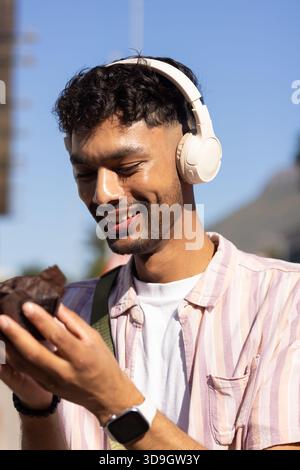 Uomo asiatico sorridente tenendo in mano un muffin al cioccolato, indossando cuffie sovrauricolari in città Foto Stock