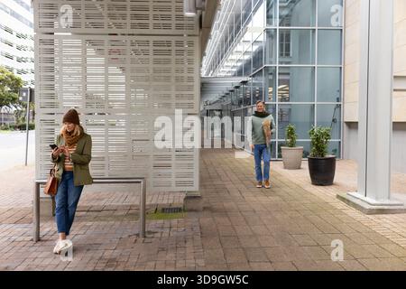 Una coppia anziana che aspetta al riparo degli autobus, controlla lo smartphone e porta con sé la borsa della spesa, copia dello spazio Foto Stock