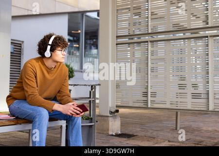 Uomo che tiene lo smartphone rosso sulla panchina in un riparo e indossa un maglione con le cuffie di notebook impilati Foto Stock