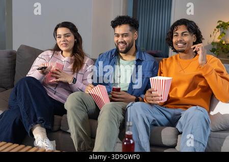 Diversi amici seduti su un divano grigio nel soggiorno che regala popcorn e bibita rossa sorseggiando a distanza Foto Stock