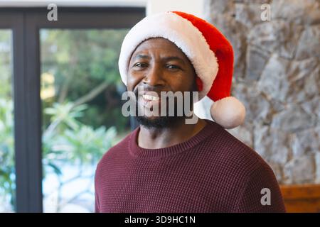 Uomo afroamericano che indossa un maglione Borgogna e un cappello di Babbo Natale davanti a una porta di vetro che mostra la piscina con il fogliame Foto Stock