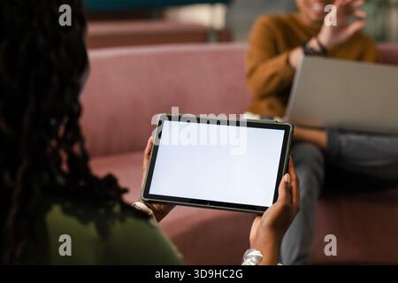 Il tablet viene tenuto su un divano rosa nella sala, con schermo bianco vuoto e un notebook sfocato Foto Stock