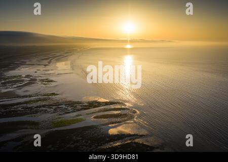 Un tranquillo paesaggio marino al tramonto presenta un orizzonte luminoso, onde dolci e piscine di marea riflettenti lungo la riva. La luce calda bagna l'acqua e l'acqua Foto Stock