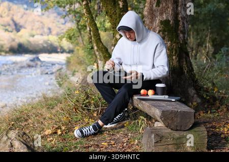 La persona con cappuccio bianco siede su un tronco di legno per albero, vibra il cibo da un contenitore nero, con un computer portatile, mele e una tazza di caffè nelle vicinanze nella foresta Foto Stock