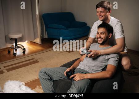 Coppie gay diverse di uomini che condividono un'accogliente serata di gioco a casa, una seduta con controller wireless, mentre il partner si appoggia per unirsi, sorridere e. Foto Stock