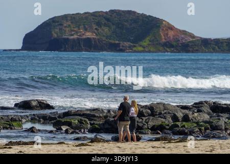 Melbourne, Victoria Australia. 6 settembre 2025. - Una coppia si trova a piedi nudi su una spiaggia sabbiosa tra rocce scure, a guardare le onde infrangersi a Bushranger Bay con spirito Foto Stock