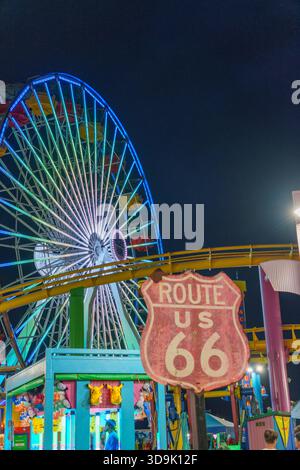 Santa Monica, CALIFORNIA, Stati Uniti. 26 settembre 2025. - Una ruota panoramica illuminata gira contro il cielo notturno, con un cartello della Route 66 in primo piano Foto Stock