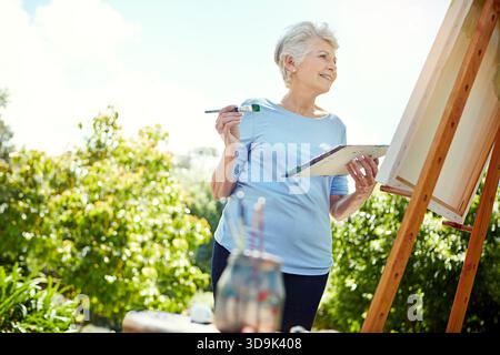 Pittura, tela e sorriso con una donna anziana in natura per progetti creativi, ispirazione e disegno del giardino. Pensionamento, artista e cavalletto con senior Foto Stock
