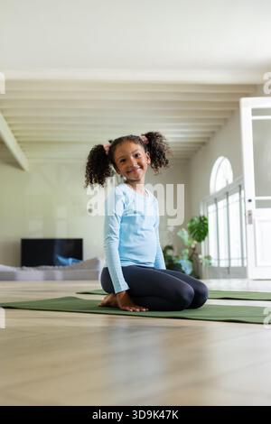Ragazza afro-americana inginocchiata su un tappetino da yoga verde nel soggiorno con abbigliamento sportivo Foto Stock