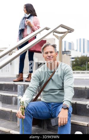 La coppia senior si riposa sui gradini urbani con un uomo che tiene margherite e smartphone mentre sorseggia un caffè Foto Stock