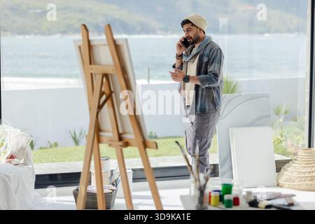 Uomo indiano in piedi in uno studio d'arte di fronte alla spiaggia che parla su smartphone e tiene in mano tazza, spazio per copiare Foto Stock