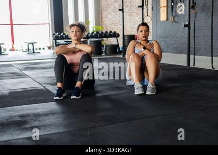 Diverse partner di allenamento femminile che eseguono sit-up su piastrelle in gomma da palestra vicino a cremagliera e anelli Foto Stock