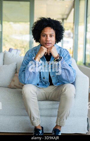 Uomo afroamericano appoggiato in avanti su un divano grigio accanto a pannelli di vetro nel soggiorno Foto Stock