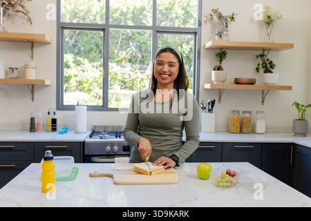 Donna che affetta panino su un piano di marmo nella cucina di casa con tagliere e contenitori di frutta Foto Stock