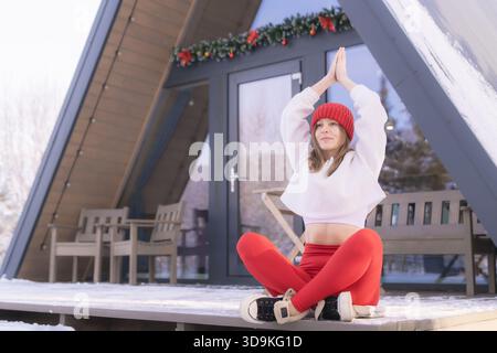 Pratica yoga invernale all'aperto con una donna che indossa un abbigliamento accogliente in un tranquillo paesaggio innevato vicino a una casa di cornici Foto Stock