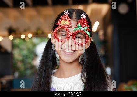 Bambina indiana sorridente mentre indossa un maglione color crema e occhiali di Natale rossi scintillanti nel caffè Foto Stock