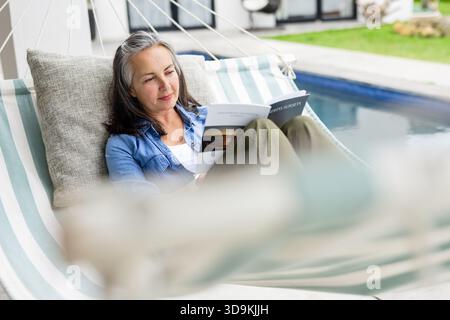 Donna anziana in camicia in denim reclinata su amaca a righe accanto alla piscina su un libro in brossura da lettura sul prato Foto Stock
