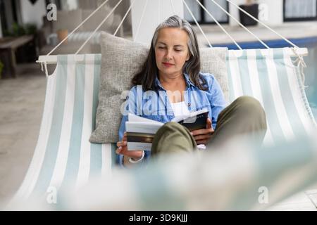 Donna anziana che si rilassa su un'amaca a righe con cuscino e libro da lettura sulla terrazza della piscina piastrellata Foto Stock
