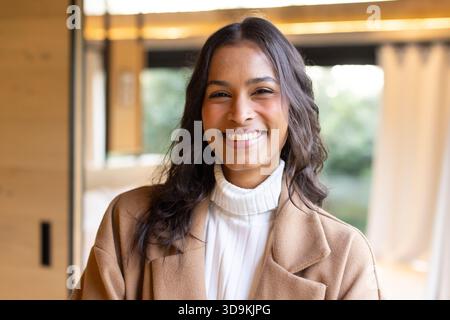 Donna che indossa dolcevita bianco e cappotto beige mentre sorride nella sala accanto alla finestra mostrando fogliame Foto Stock