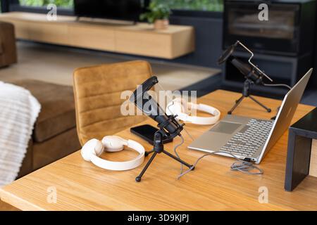 La configurazione del podcast mostra i microfoni del condensatore, le cuffie, il computer portatile e lo smartphone sul tavolo in salotto Foto Stock
