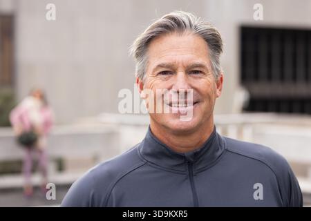 Uomo di mezza età sorridente alla fotocamera mentre si trova all'aperto in Urban plaza con una giacca grigio scuro Foto Stock