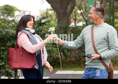 Coppia senior che scambia fiori bianchi sul percorso lastricato del parco tenendo in mano smartphone e borsa tote rossa Foto Stock