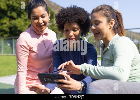 Diverse amiche di abbigliamento sportivo appoggiate sulla panchina al tablet che si sta avvicinando al parco indossando uno smartwatch Foto Stock