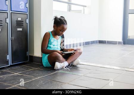 Ragazza afroamericana seduta nella sala della scuola usando un tablet con scarpe da ginnastica turchesi con armadietti Foto Stock