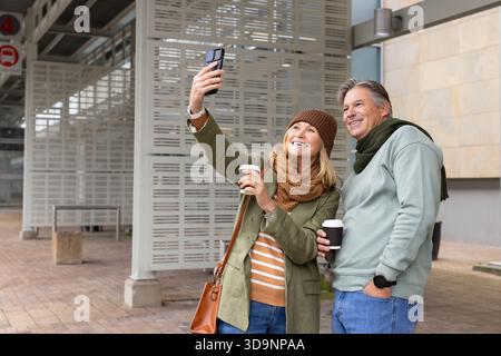 Coppia senior che tiene in mano tazze da caffè e scatta selfie con lo smartphone sulla piattaforma al segnale di fermata dell'autobus Foto Stock