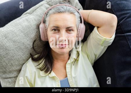 Donna anziana sdraiata sul divano di casa, che riposa la testa su un cuscino grigio e indossa cuffie dorate Foto Stock