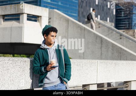 Uomo afro-americano appoggiato alla barriera del plaza che tiene una tazza di caffè e uno smartphone, spazio per copiare Foto Stock