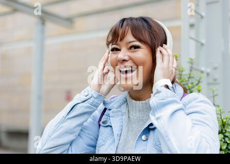 Donna adulta con cuffie bianche e sorridente sotto la cornice metallica all'esterno con smartwatch Foto Stock