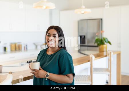 Donna afroamericana che tiene in mano una tazza di ceramica bianca e sorride sull'isola della cucina sotto le luci a sospensione Foto Stock
