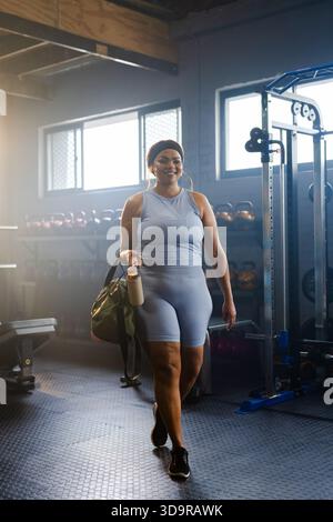 Donna di mezza età che cammina verso la macchina fotografica in palestra in abbigliamento sportivo con borsone verde e bottiglia beige Foto Stock