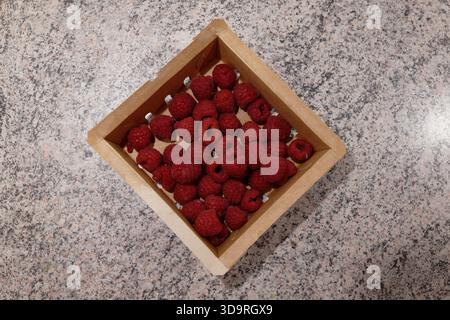 Un piccolo contenitore di carta pieno di lamponi rossi freschi si trova su una superficie del tavolo in grafite grigio scuro, che mostra un semplice spuntino naturale. Foto Stock